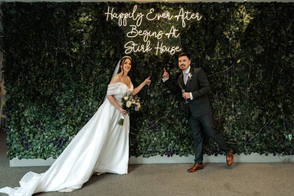 Bride and groom posing next to Happily Ever After Begins At Stirk House neon sign on foliage wall.
