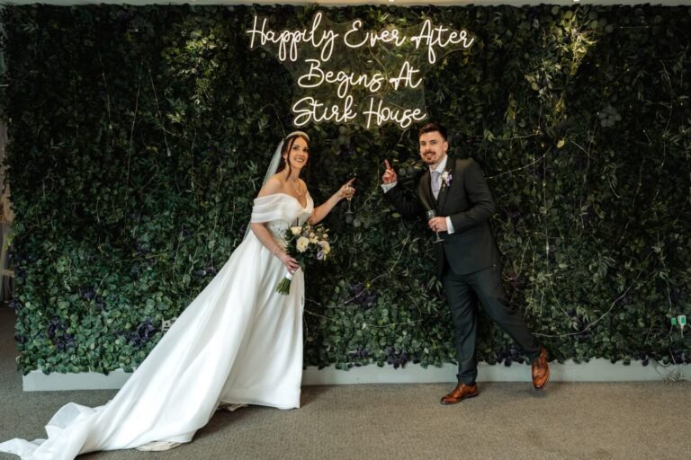 Bride and groom posing next to Happily Ever After Begins At Stirk House neon sign on foliage wall.