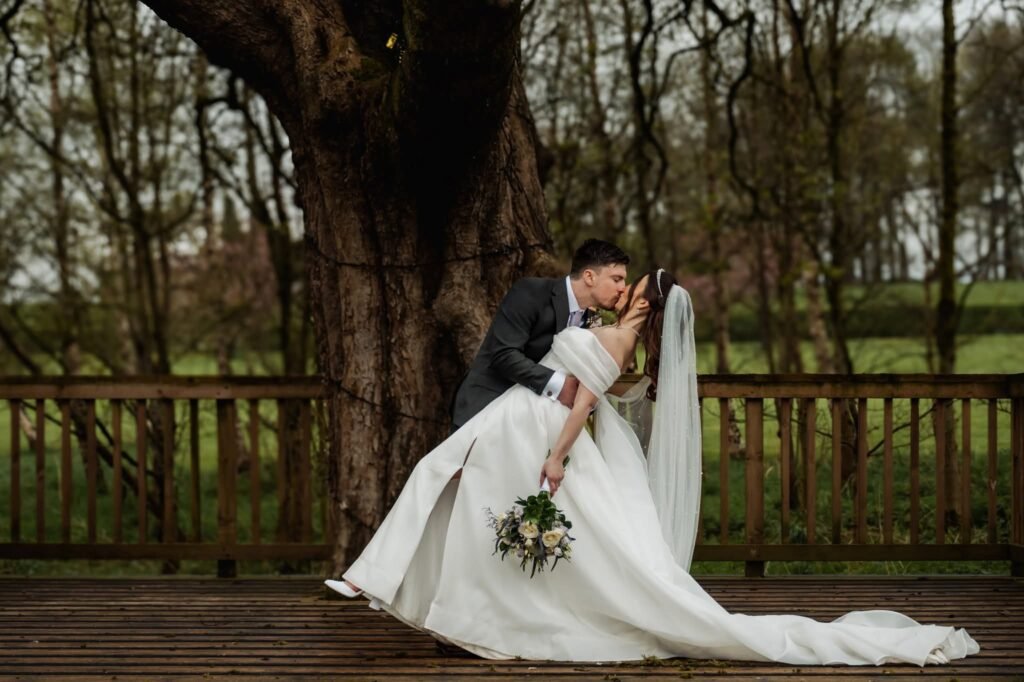 Romantic groom dipping bride for a kiss on the decking at Stirk House with rustic woodland background.