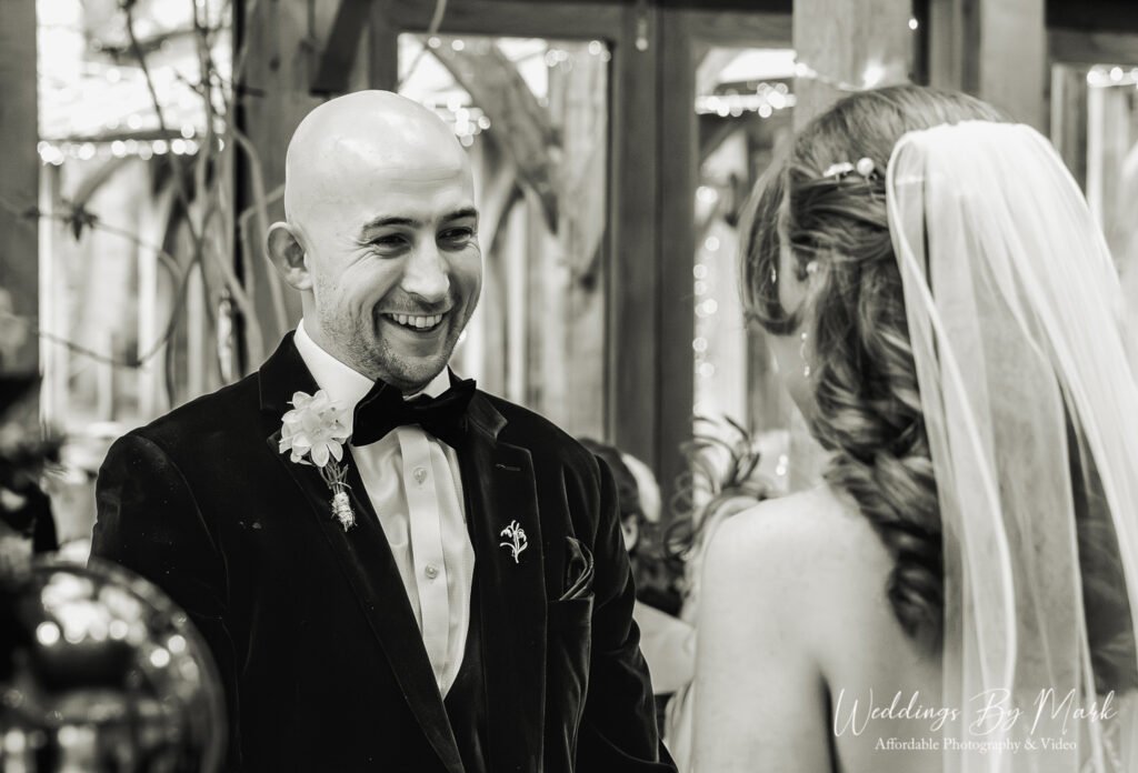 Black and white documentary wedding photography showing Mathew smiling at his bride during vows at The Oak Tree of Peover