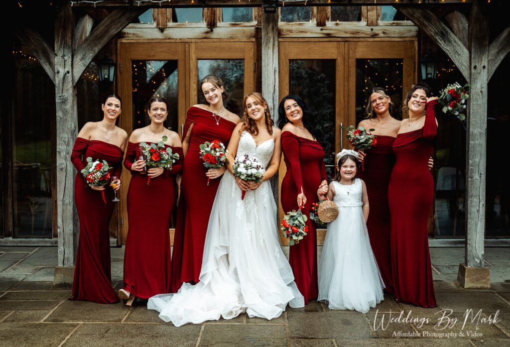 Bride Katie posing with bridesmaids in dark red dresses outside the rustic barn doors at The Oak Tree of Peover