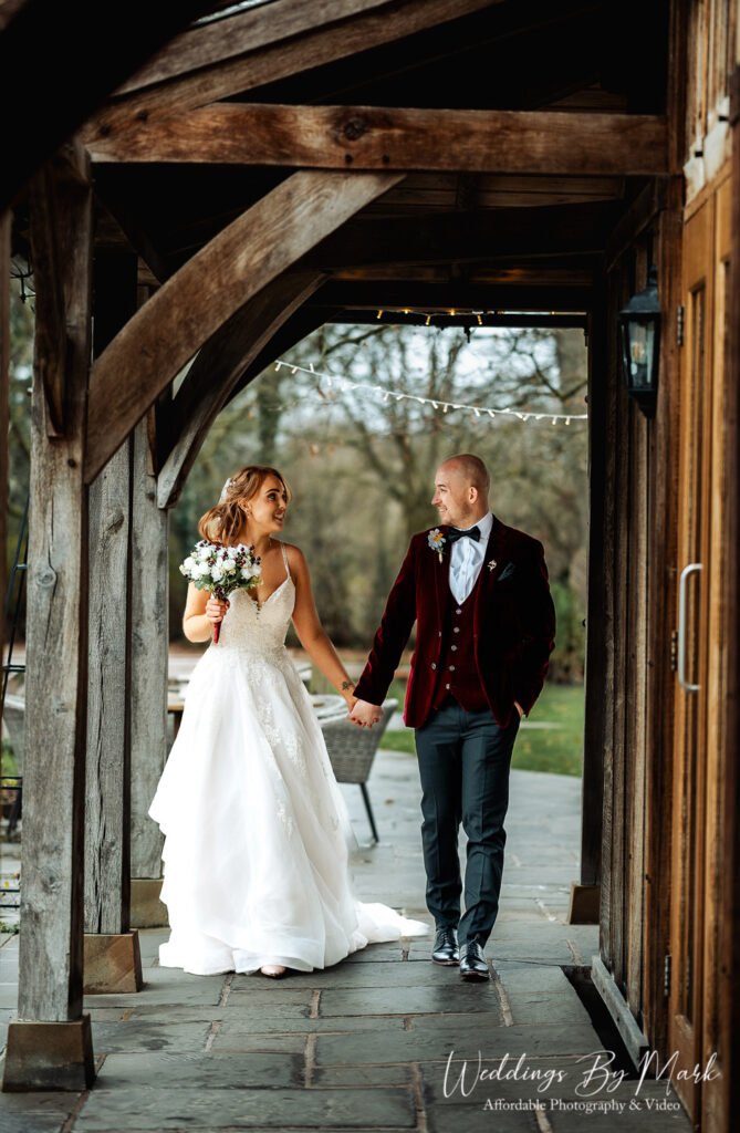 Katie and Mathew walking hand-in-hand under the rustic timber entrance at The Oak Tree of Peover during their couple portraits.