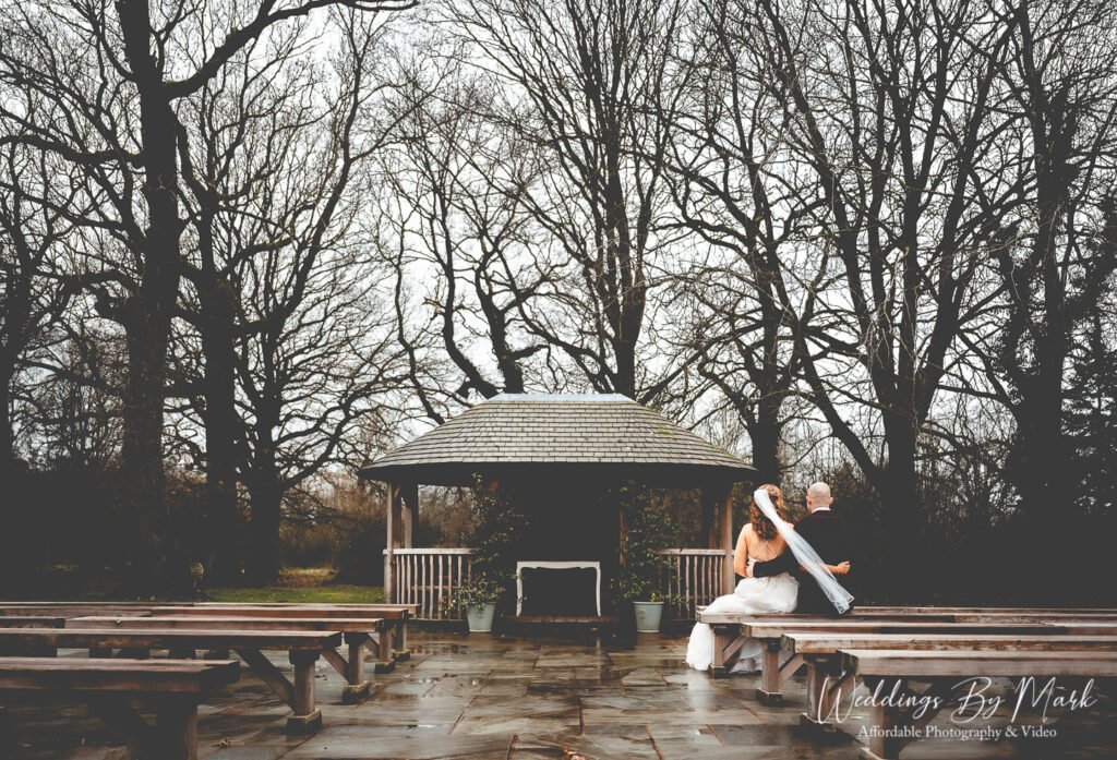 Wide scenic shot of the newlyweds sitting on a bench overlooking the gazebo on the grounds of The Oak Tree of Peover
