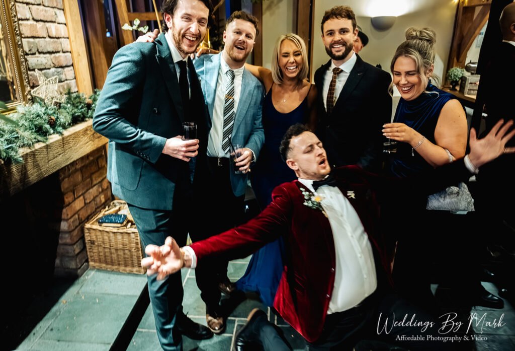 A fantastic, high-energy party shot showing the groom doing a rockstar knee slide on the floor while his friends cheer him on from behind