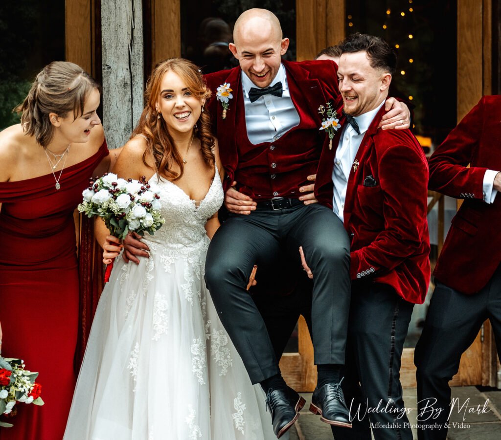 Fun wedding photography moment showing groomsmen lifting Mathew while Katie laughs outside The Oak Tree of Peover