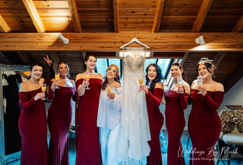Bridesmaids in dark red dresses toasting champagne with Katie during bridal prep at The Oak Tree of Peover