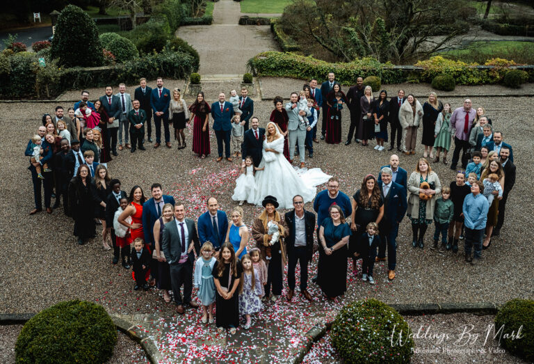 Maria and Jonny surrounded by guests in a heart shape at their Eaves Hall wedding in Lancashire.
