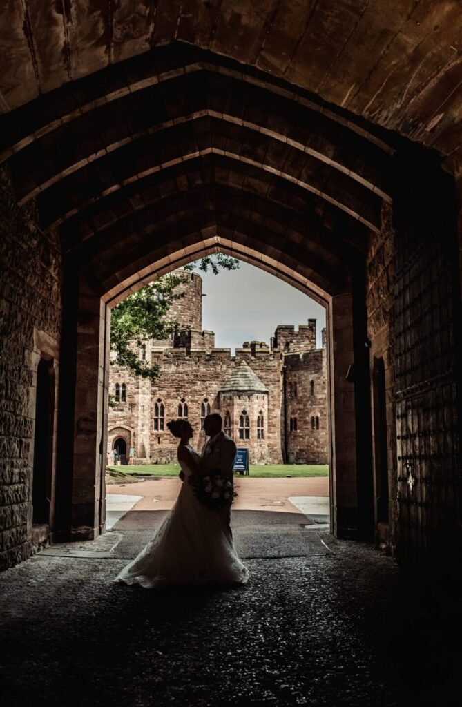Dramatic silhouette wedding portrait of Jade and Nick framed by the grand stone entrance archway at Peckforton Castle.