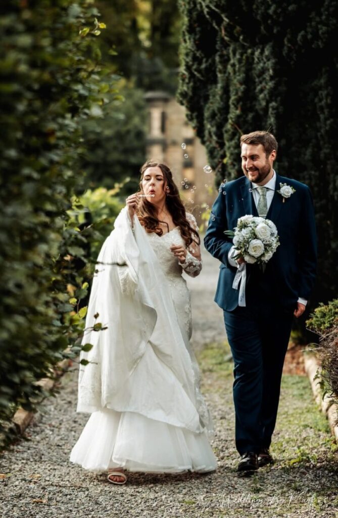 Bride blowing bubbles as the groom smiles beside her on a garden path at Ringwood Hall