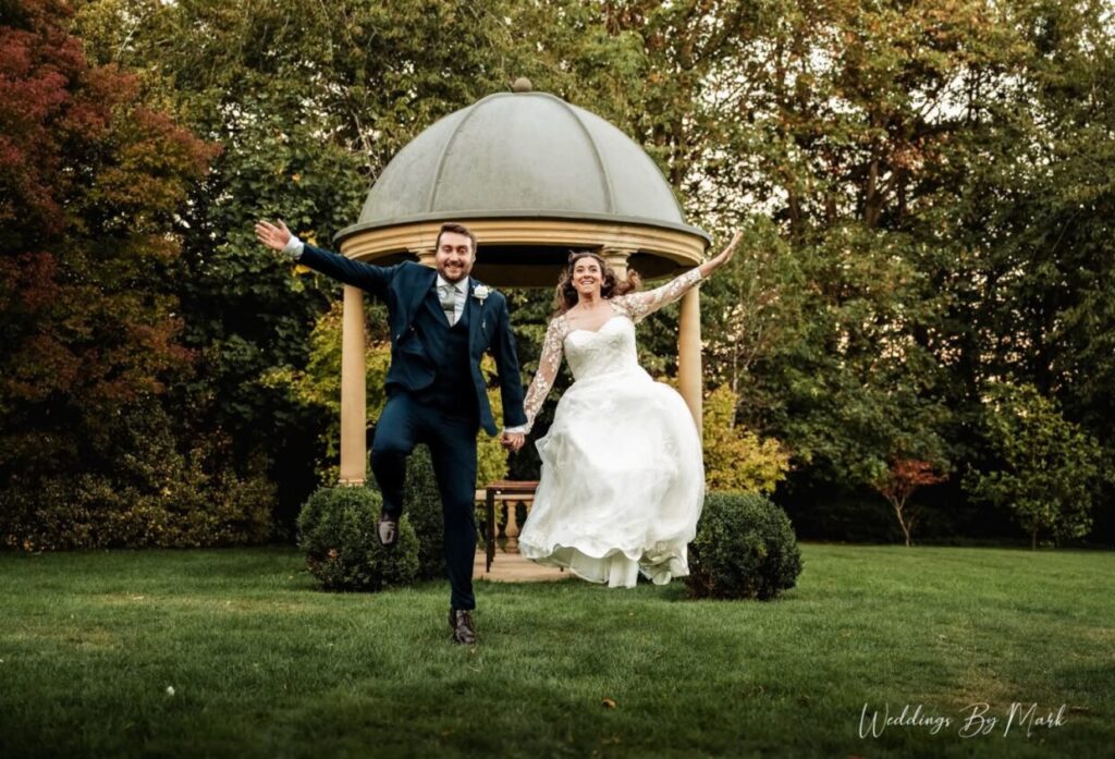 Bride and groom jumping together by a garden gazebo at Ringwood Hall Hotel and Spa
